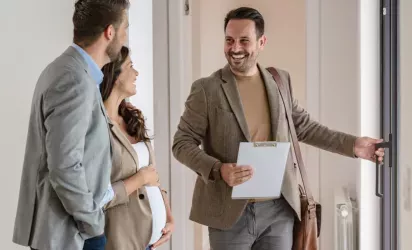A real estate agent smiling while opening a door for a couple, with the woman appearing pregnant, during a property showing.