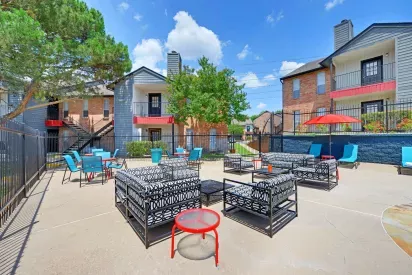 Vibrant outdoor community space featuring stylish seating areas with modern patio furniture, surrounded by colorful apartment buildings and lush greenery. The bright blue sky and open layout create an inviting and lively atmosphere for social gatherings.