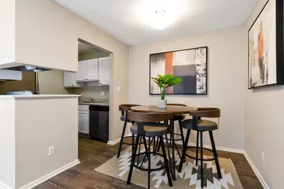 A cozy dining area with a round wooden table, modern chairs, and abstract artwork, adjacent to a compact kitchen with white cabinetry.