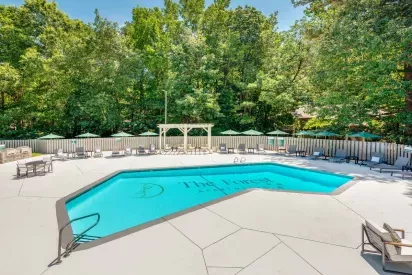 A bright outdoor pool area surrounded by lounge chairs, green umbrellas, and a shaded pergola, located at The Forest Apartments, with a backdrop of lush greenery.