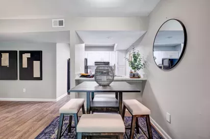 A modern dining area featuring a wooden table with cushioned stools, a decorative centerpiece, and a view into a bright kitchen.