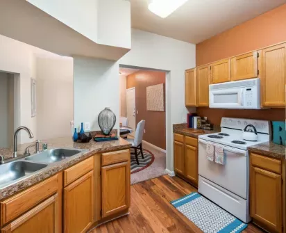 Cozy and modern kitchen featuring warm wooden cabinetry, granite countertops, and white appliances. The open layout includes a double sink, stylish decor accents, and hardwood-style flooring that transitions into a dining area with a contemporary design, perfect for comfortable and functional living.