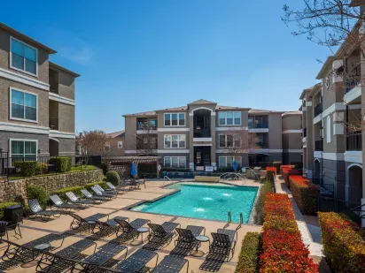 A bright courtyard with a sparkling pool, lined with lounge chairs, red shrubs, and modern apartment buildings in the background.