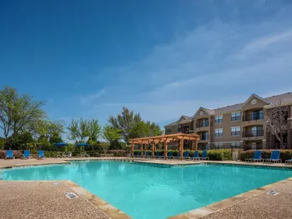 A sparkling outdoor pool surrounded by lounge chairs and a pergola, with apartment buildings and lush greenery in the background under a clear blue sky.