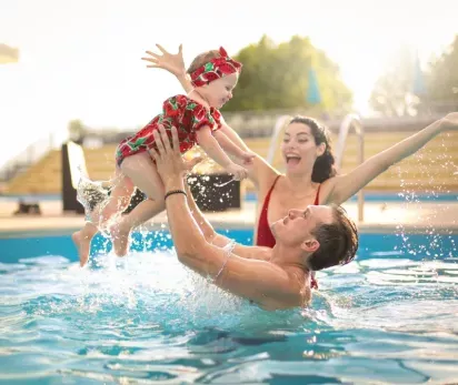 Joyful family enjoying a sunny day at the pool, with a father playfully lifting a smiling baby in the air while the mother cheers with excitement. The sparkling water and warm, vibrant setting create a fun and inviting atmosphere perfect for leisure and family bonding.