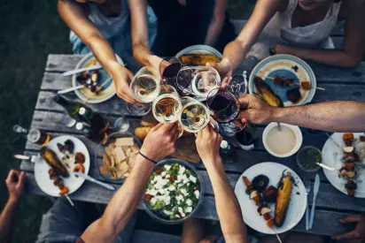 A group of people raising glasses in a toast during a festive outdoor meal with a variety of food on a wooden table.