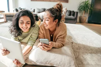 Two friends lounging on cushions in a cozy living room, smiling and engaging with a tablet and a smartphone.