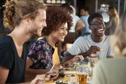 A group of people sitting at a table, smiling and enjoying drinks in a restaurant setting, with food and tableware present.