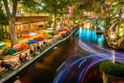 A scenic view of a vibrant riverside dining area illuminated with colorful lights and adorned with bright umbrellas, showcasing the energy and charm of a waterfront destination.