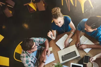 A group of young professionals collaborating around a wooden table in a vibrant, modern café, with a laptop, notebooks, and coffee cups, creating a warm and productive atmosphere.