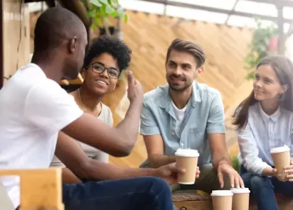 A group of diverse friends enjoying a casual conversation over coffee in a cozy, outdoor café setting, surrounded by natural wooden décor and greenery.