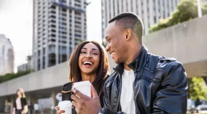 A man and woman are outdoors on a city street, laughing together. Both are smiling and appear to be enjoying a light-hearted moment while interacting with each other. The woman is holding a cellphone. They are dressed casually, with buildings in the background.