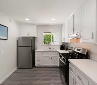Modern kitchen with sleek white cabinetry, stainless steel appliances, and a gas stove. Features a spacious countertop, bright natural light from a window above the sink, and wood-style flooring for a clean, contemporary look.