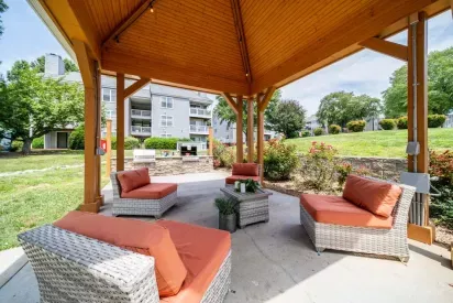 An outdoor pavilion with comfortable wicker seating featuring orange cushions, overlooking landscaped greenery and apartment buildings.