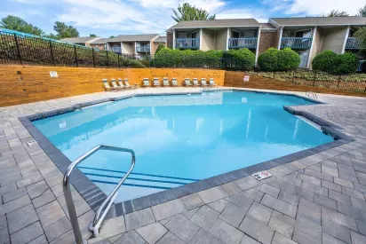An outdoor swimming pool surrounded by a brick-paved deck, lounge chairs, and apartment buildings in the background.