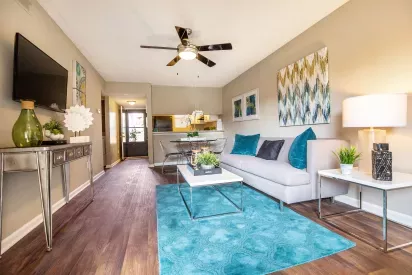 A modern living room with a bright teal rug, stylish furniture, wall art, and an open view into the kitchen and dining area.
