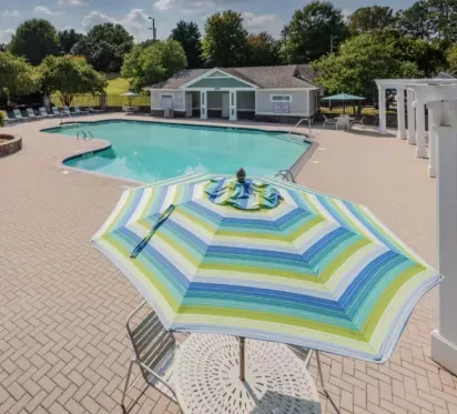 A sparkling outdoor pool surrounded by lounge chairs, with a colorful striped umbrella shading a round table on a brick-paved patio.