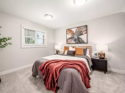 A cozy bedroom with a neatly made bed adorned with throw pillows and a red blanket, flanked by nightstands and illuminated by soft lighting.