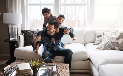 A joyful father enjoying quality time with his two sons, giving them piggyback rides in a cozy living room. The trio shares a fun and laughter-filled moment, creating lasting memories in a warm, comfortable home environment.