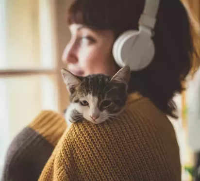 A young woman wearing headphones gazes out of a window while a relaxed kitten rests on her shoulder, wrapped in the warmth of her cozy sweater.