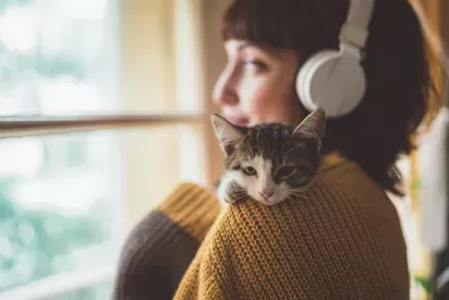 A woman wearing headphones gazes out a window while a kitten rests on her shoulder, nestled against her yellow sweater.