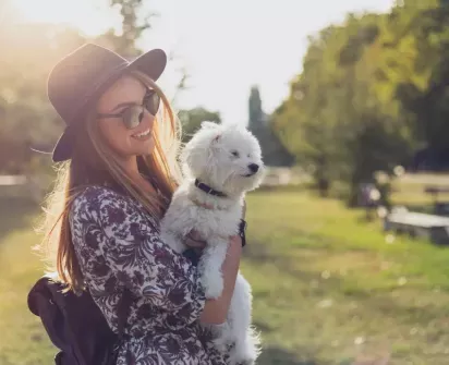 A woman is smiling while holding a small white dog in her arms, set against a sunlit park background. She is wearing a hat and sunglasses.