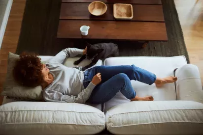 A woman lounging on a white couch is petting her black dog, with a wooden coffee table and cozy decor in the background.