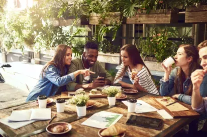 A group of young friends enjoying a casual meeting at a cozy outdoor cafe, sharing food, drinks, and laughter. Surrounded by greenery, they're engaged in conversation while having light snacks and coffee.
