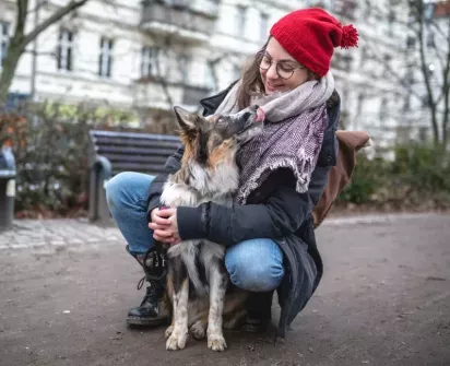  Woman smiling and embracing her dog outdoors in a park setting, bundled up in a cozy red hat and scarf, sharing a joyful and affectionate moment.