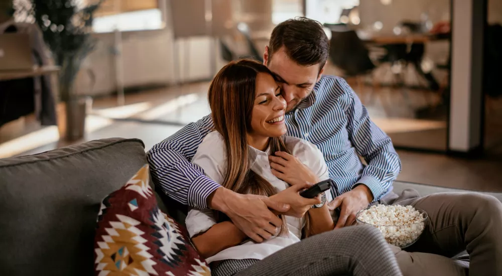 A couple sits closely on a sofa, smiling and enjoying a bowl of popcorn while holding a remote control.