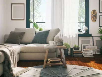 A cozy living room with a light beige sectional sofa, a wooden coffee table, and abundant natural light streaming through large windows adorned with sheer white curtains.