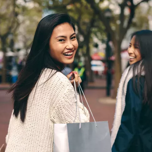Two smiling friends carrying shopping bags while walking outdoors in an urban setting.