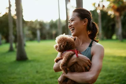 A smiling woman enjoys a sunny day at the park, holding her fluffy brown dog close.