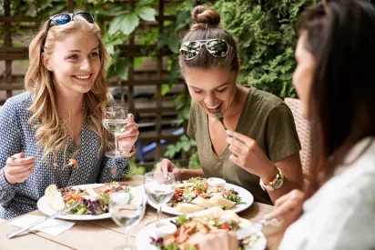  A group of three women enjoying a lunch together outdoors, sharing laughs as they eat fresh salads and drink water in a sunny, leafy setting. The relaxed atmosphere captures the joy of good food and even better company.
