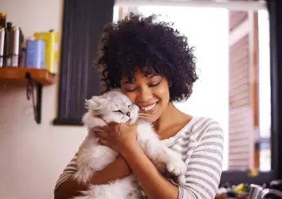 A smiling woman cuddles a fluffy gray cat in a cozy kitchen setting.