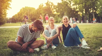 Three friends sit together on the grass in a sunny park, laughing and enjoying a relaxed moment surrounded by trees and people in the background.