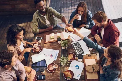 A group of diverse colleagues share pastries and coffee while collaborating on documents and using a laptop at an outdoor table.
