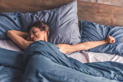 A woman wakes up smiling in bed, stretching her arms under cozy blue bedding in a sunlit bedroom.
