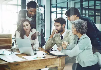 A diverse group of five professionals collaborates around a wooden table in a bright, modern office, focusing intently on a laptop screen during a discussion.
