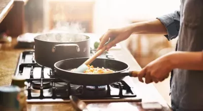 A cozy kitchen scene featuring a person stirring vegetables in a frying pan while another pot steams on the stovetop, illuminated by warm natural light.