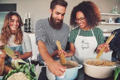 A smiling couple prepares pasta together in a kitchen, with the woman wearing an apron that says "Be my Guest."