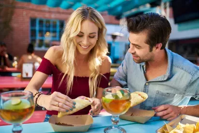 A couple enjoying a casual, sunny lunch together at a colorful outdoor restaurant, savoring tacos and drinks while sharing a lighthearted moment. The vibrant atmosphere adds to the joy of their time together.