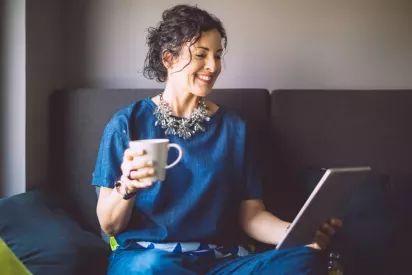 A smiling woman relaxes on a sofa, holding a cup of coffee and looking at a tablet.