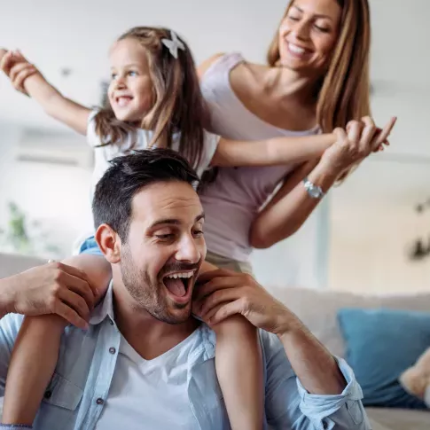 A family happily playing together at home, with the father carrying the child on his shoulders while the mother smiles alongside them.