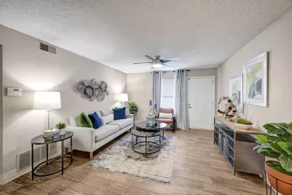 A cozy living room with a modern design, featuring a sofa, decorative artwork, and a ceiling fan, complemented by wood-style flooring and soft lighting.
