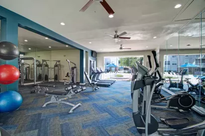 A spacious fitness center with cardio machines, weight equipment, exercise balls, and large mirrors, featuring bright blue flooring and views of an outdoor pool area through the windows.