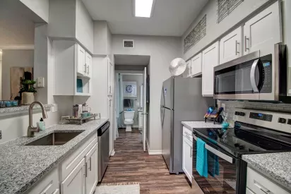 A modern galley kitchen with sleek white cabinets, granite countertops, stainless steel appliances, and decorative teal accents, leading to a bathroom in the background.