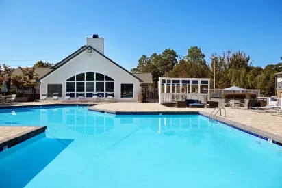 An outdoor pool area with crystal-clear water, surrounded by a lounge area, white pergola, and a clubhouse in the background.