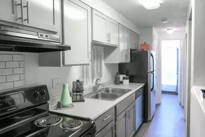 A modern galley kitchen with white cabinets, black appliances, a double sink, and a sleek countertop, leading to a well-lit hallway.