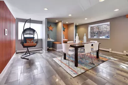 A stylish communal space featuring a modern dining table with white chairs, a colorful rug, cozy computer work nooks, and a unique hanging chair with a playful orange cushion.